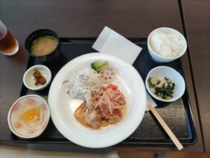 クロスウェーブ府中の昼食 中央にサラダとドレッシングをかけた鶏の唐揚 右にシラスのわかめ和え、右上に白ごはん、左上に味噌汁、その下に漬物、左下に、フルーツゼリー